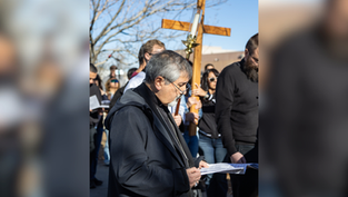Hombre de negro lee un papel en una reunión religiosa al aire libre, con una cruz de madera y gente al fondo.