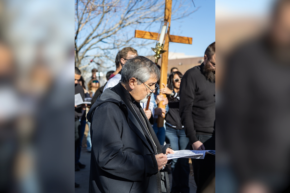 Hombre de negro lee un papel en una reunión religiosa al aire libre, con una cruz de madera y gente al fondo.