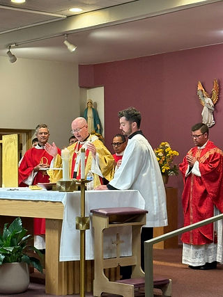 Sacerdotes celebran misa ante el altar con velas; uno reza, otro lee, en capilla con pared rosa y estatua religiosa.