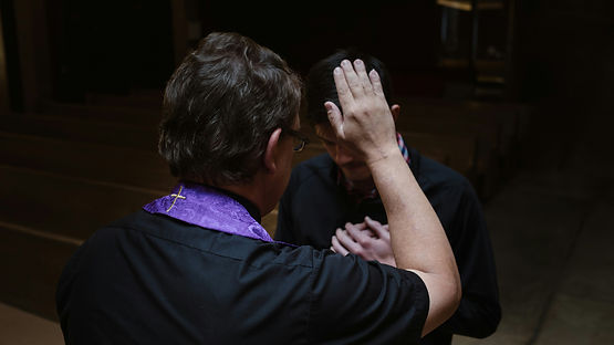 Un sacerdote con estola morada bendice a un hombre en actitud reflexiva en una iglesia oscura. Ambiente solemne.