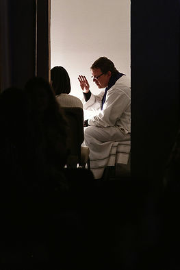 Un hombre con ropa blanca levanta la mano hacia una persona sentada frente a él en una sala iluminada, creando un ambiente solemne.