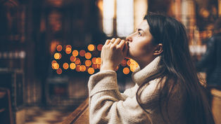 Mujer pensativa con suéter claro, mirando al frente en un interior cálido con luces desenfocadas y ambiente tranquilo.